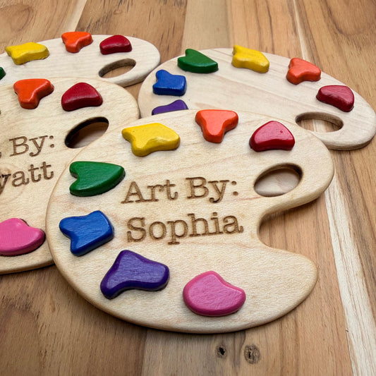 Four personalized wooden paint palette magnets with colorful paint pieces embedded in them, arranged on a wooden surface. Showing closeup of magnet with engraving 'Art By: Sophia'.