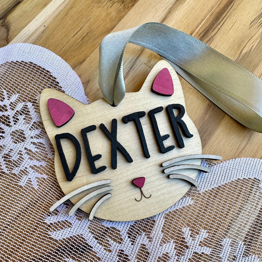 Cat face shaped ornament on maple with black lettering. Pink painted ears and nose. White painted whiskers.