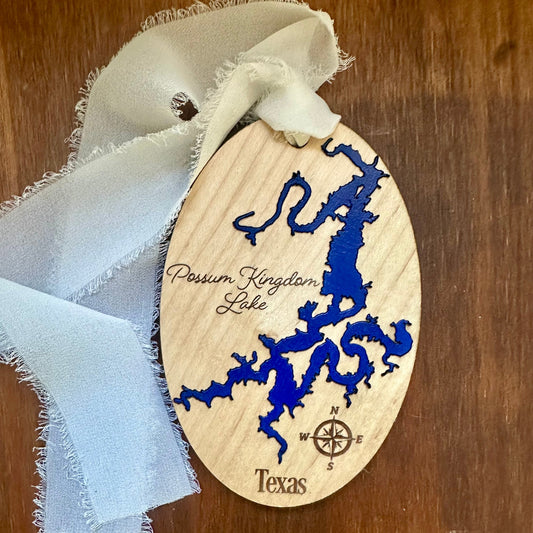 An oval wooden ornament featuring a blue laser-engraved map of Passum Kingdom Lake, Texas, with a white fabric ribbon, shown on a dark wood background.