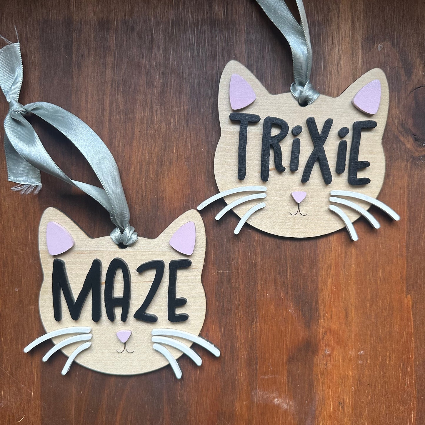 Two wooden cat-shaped ornaments with the names 'TRIXIE' and 'MAZE' in black letters, decorated with light pink ears, white whiskers, and hanging from green ribbons on a dark wood background.