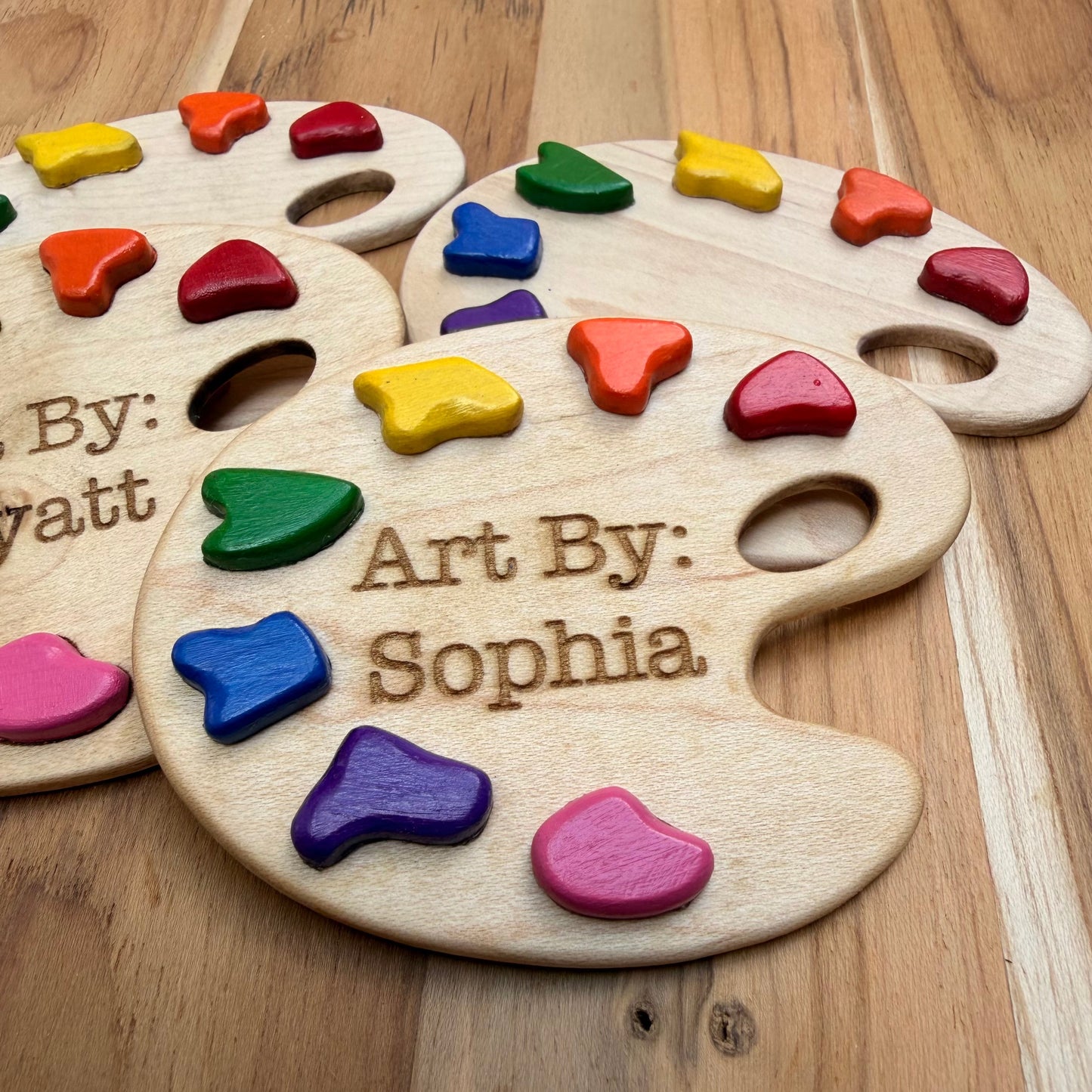 Four personalized wooden paint palette magnets with colorful paint pieces embedded in them, arranged on a wooden surface. Showing closeup of magnet with engraving 'Art By: Sophia'.