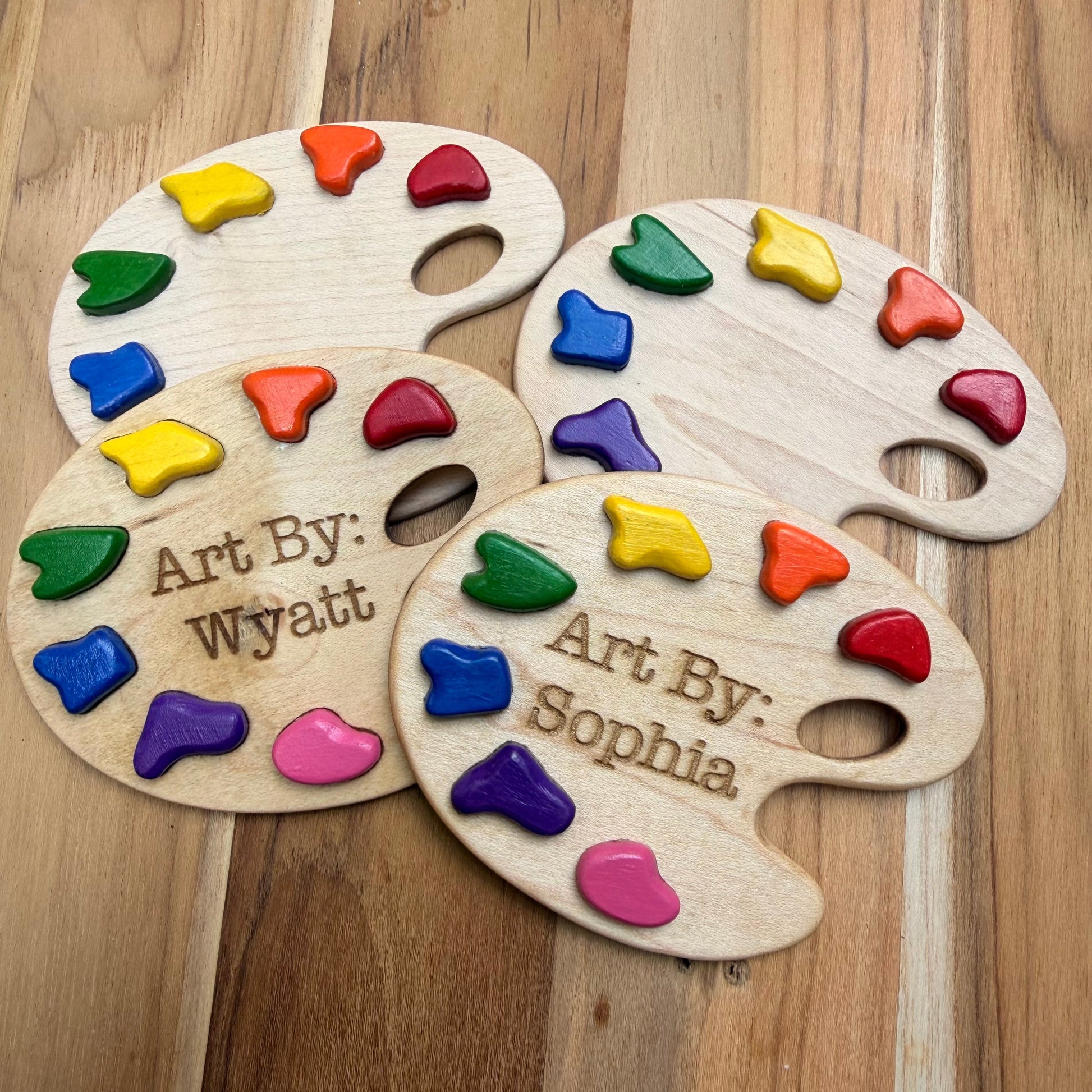 Four personalized wooden paint palette magnets with colorful paint pieces embedded in them, arranged on a wooden surface. Two palettes are engraved with names: 'Art By: Wyatt' and 'Art By: Sophia'.