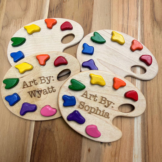 Four personalized wooden paint palette magnets with colorful paint pieces embedded in them, arranged on a wooden surface. Two palettes are engraved with names: 'Art By: Wyatt' and 'Art By: Sophia'.