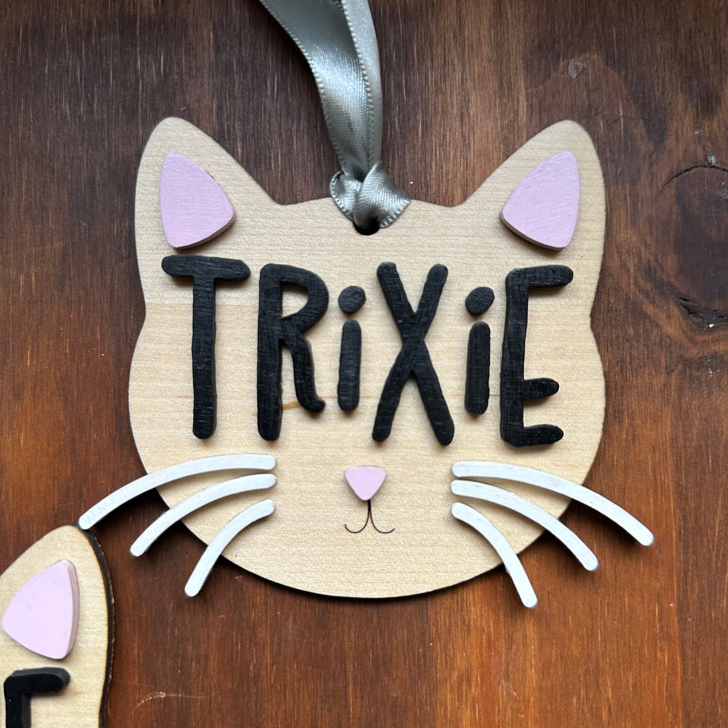 A wooden cat-shaped ornament with the name 'TRIXIE' in black letters, decorated with white whiskers, pink ears, and a small pink nose, shown on a dark wood background.