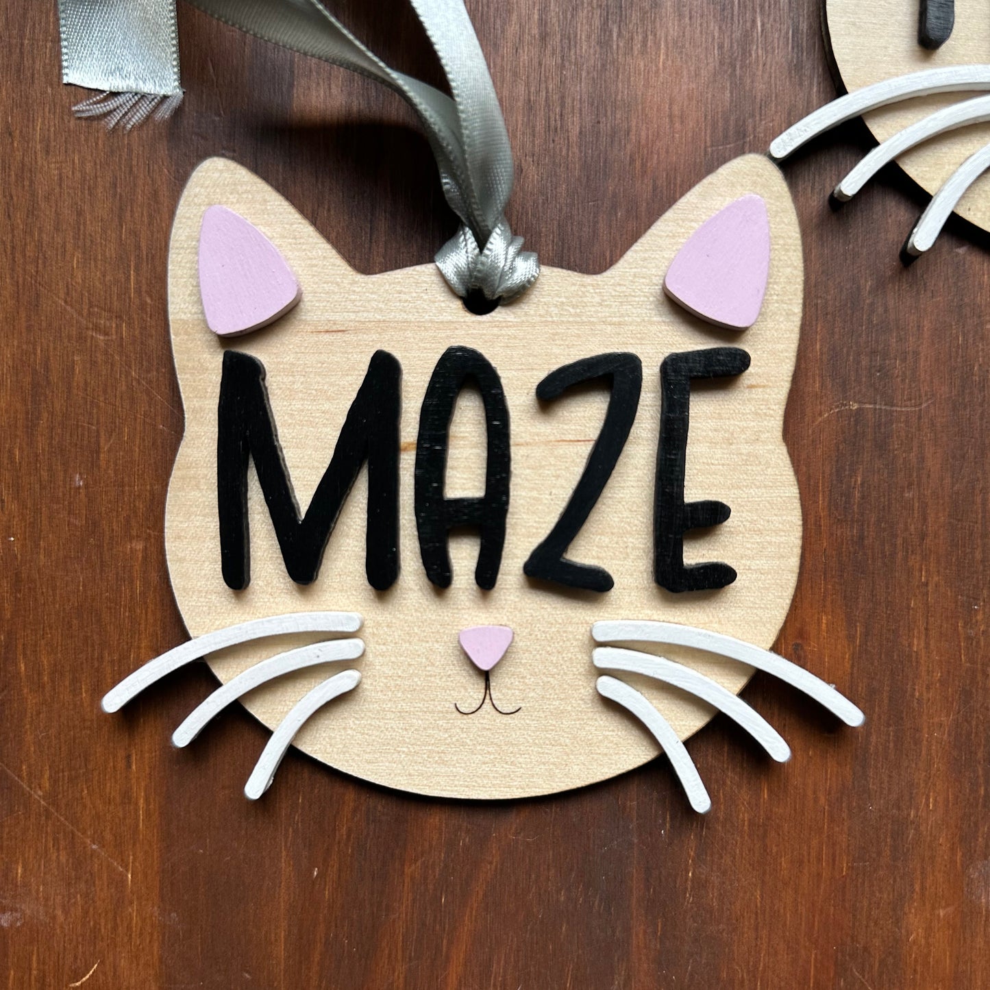 A wooden cat-shaped ornament with the word 'MAZE' in black letters, decorated with white whiskers, pink ears, and a green ribbon, placed on a dark brown wooden surface.