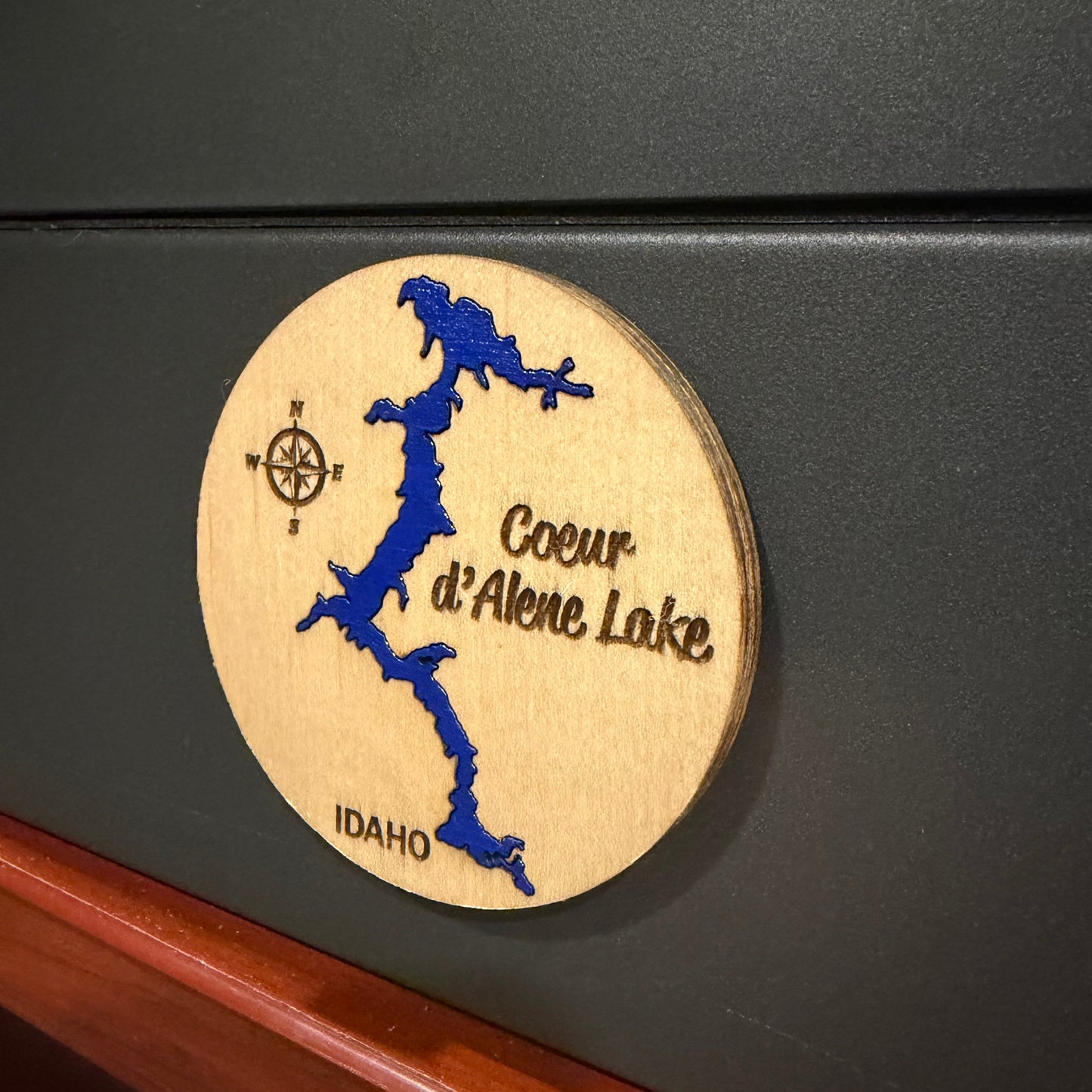 A round wooden magnet featuring a blue map design of Coeur d'Alene Lake, Idaho, with engraved text and a compass, shown magnetized a dark gray background. 