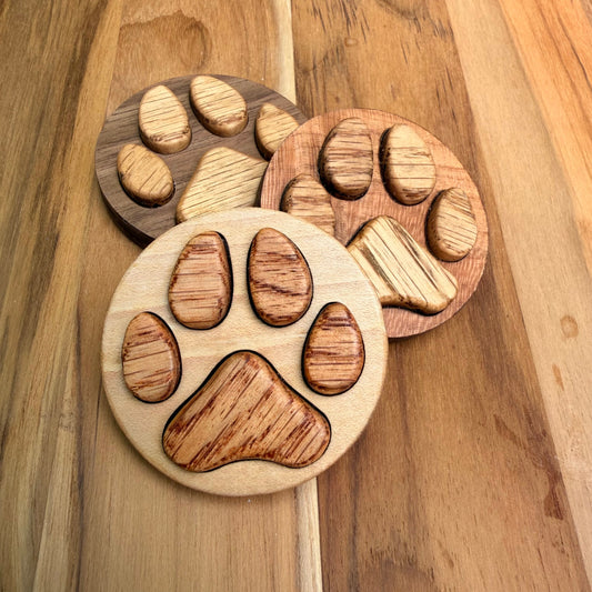 Three round wooden paw print magnets in different wood tones (one maple, one cherry, one walnut), arranged on a wooden surface.