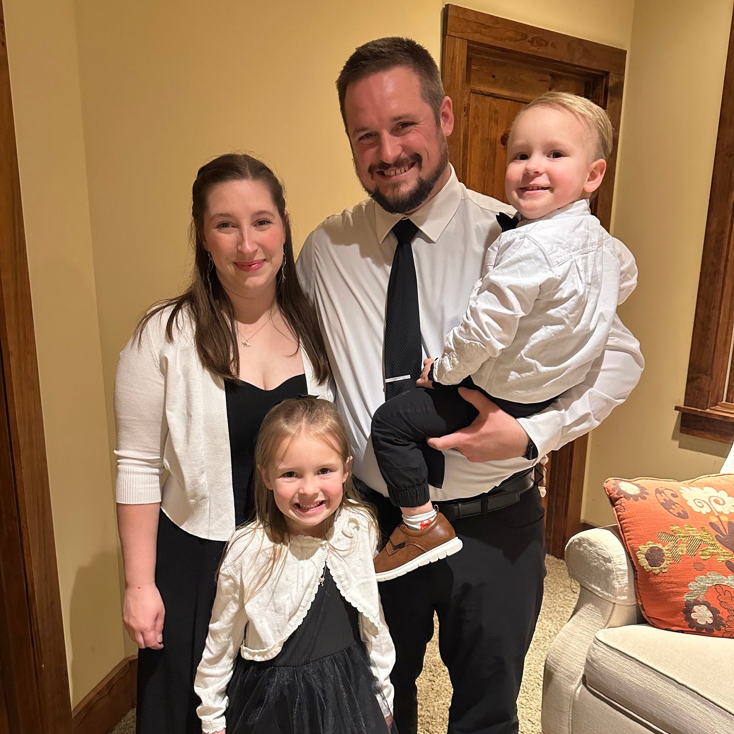 A family of four posing together in an indoor home setting. The father holds a young boy wearing a white dress shirt and black pants, while the mother and daughter wear black and white outfits.