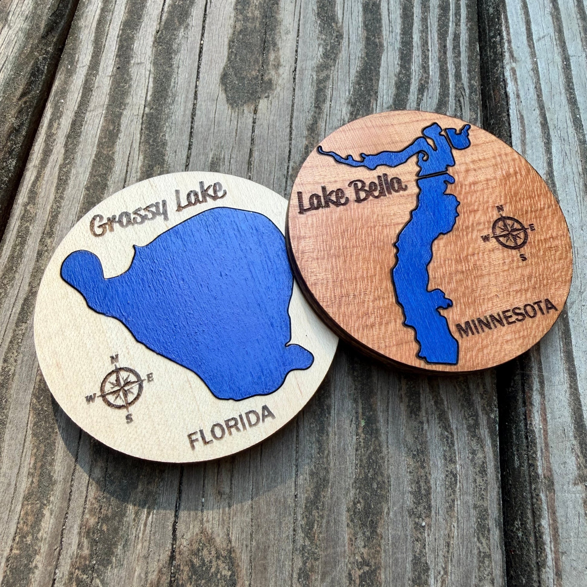 Two round wooden magnets featuring Grassy Lake, Florida and Lake Bella, Minnesota, displayed on a weathered gray wooden surface. Each magnet shows cobalt blue lake with lake name, state, and compass.