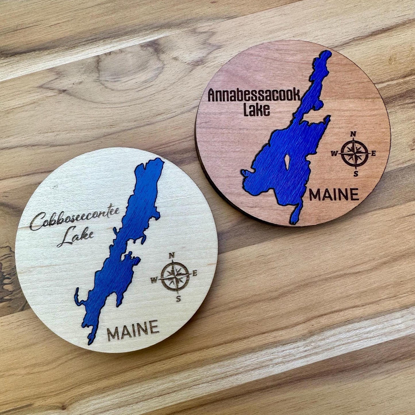 Two round wooden magnets featuring engraved maps of Maine lakes (Cobboseecontee Lake and Annabessacook Lake) with compass, lake name, and state displayed on a light wood surface. Lake is a cobalt blue. 