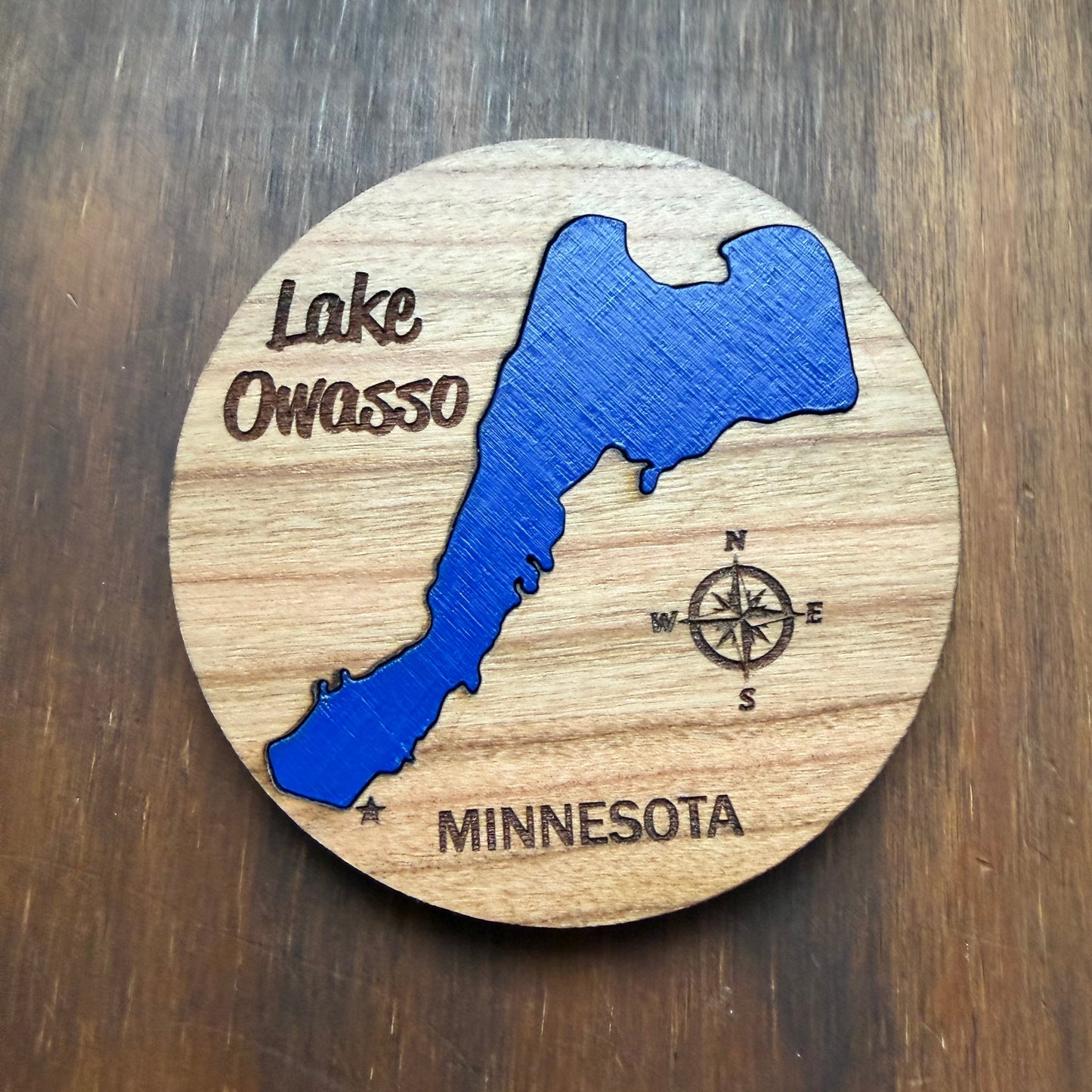 A round wooden magnet featuring a laser-engraved map of Lake Owasso, Minnesota, with a blue lake silhouette, compass, and the words 'Lake Owosso' and 'MINNESOTA' on a natural wood grain background. 