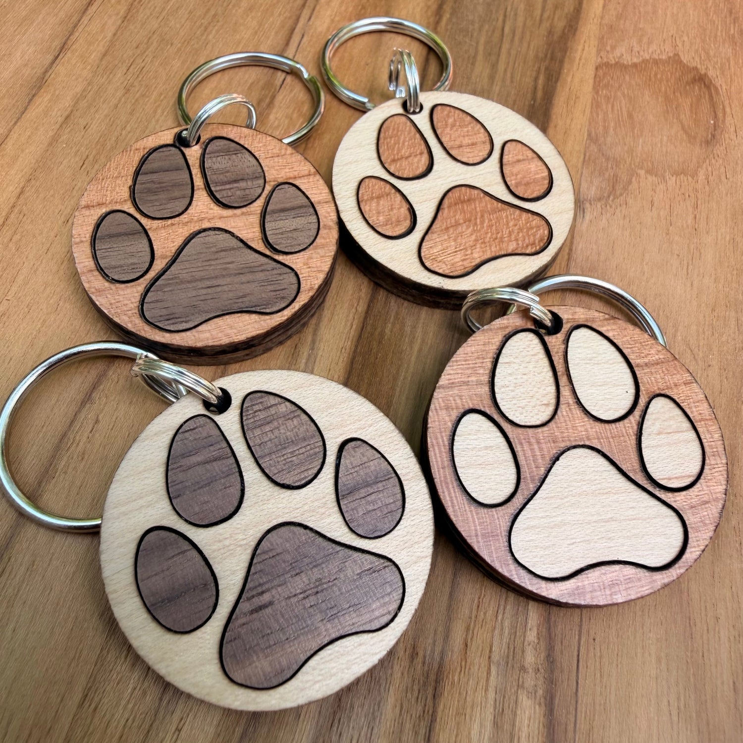 Four round wooden paw print keychains with silver metal key rings, arranged on a wooden surface.