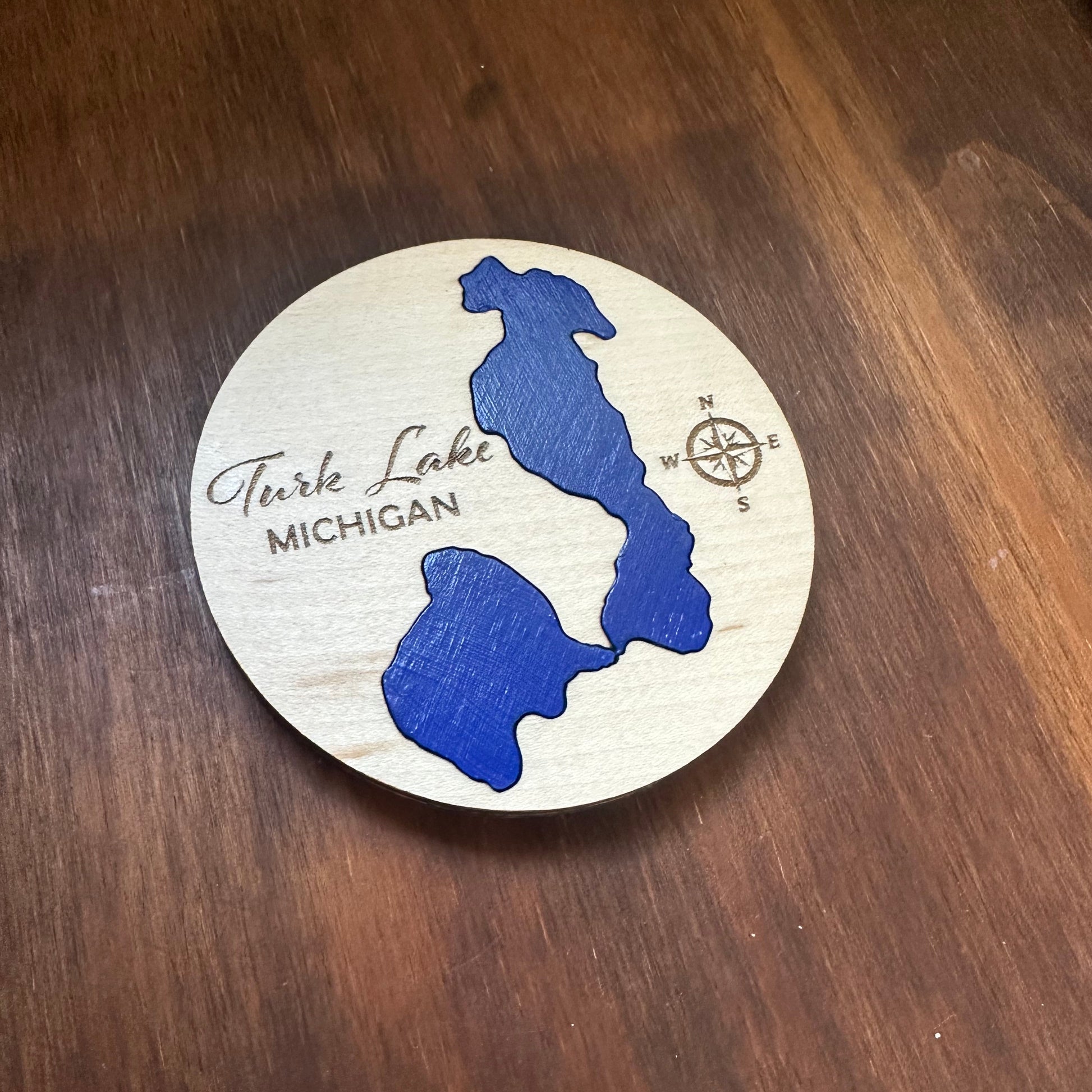 A round wooden magnet featuring a blue map design of Turk Lake, Michigan, with a compass and the words 'Turk Lake MICHIGAN' engraved on maple. Lake is a cobalt blue.